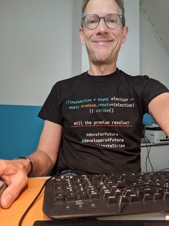 Ingo sitting at desk in front of a keyboard wearing a t-shirt with pseudo-code: climateAction = async election => await Promise.resolve(election) || strike() Will the promise resolve? #devsForFuture #developers4future climateStrike