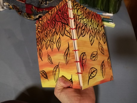 A book held to the camera showing the open spine with coptic binding.

The covers are a Mix of yellow and red with black leaves painted on
