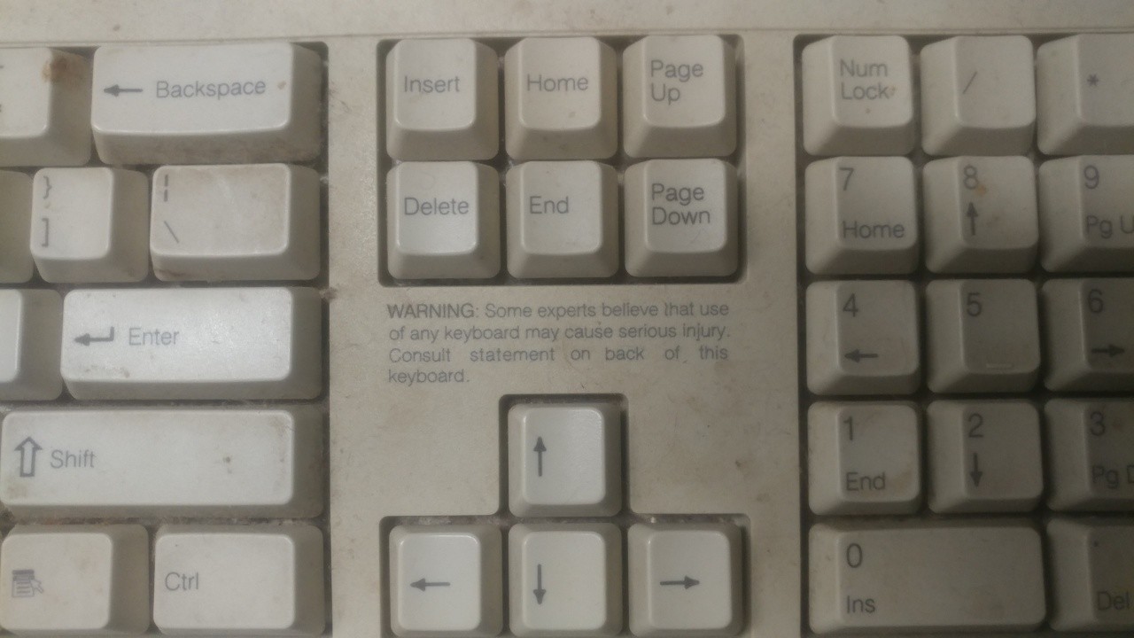 Old beige keyboard with a warning above the arrow keys. It says "Warning: some experts believe that use of any keyboard may cause serious injury."