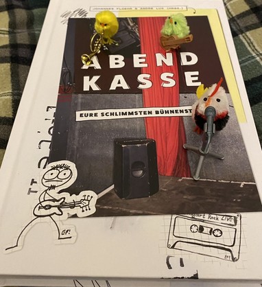 Drei Küken mit verschiedenen Instrumenten und Mikrofon, stehen auf dem Buch "Abendkasse", in dem es um schlimme Bühnenstorys geht.