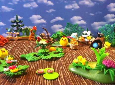 Küken bei der Ostereiersuche in der Natur. Der Boden ist braun, überall sind grüne Pflanzen und Bäume, in denen Eier versteckt sind. Im Hintergrund ein Hase mit Bollerwagen in denen bunte Eier liegen. Der Himmel im Hintergrund ist blau mit Wolken.