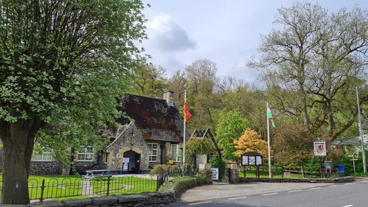 Picture of a stone built village hall in a pretty setting of green grass and circular paths. Surrounded by trees and hills. Two flags are flying. One is the flag of Owain Glyndwr and has predominant colours of red and gold. The other flag is the Welsh flag. White and green with a red dragon. The hall has posters on the gate and door stating it is a polling station.