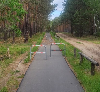 Ein Radweg durch den Wald, schmal, aber gut asphaltiert, dann plötzlich kurz hintereinander zwei enge Drängelgitter.
Parallel zum Radweg verläuft ein sandiger Waldweg. 
