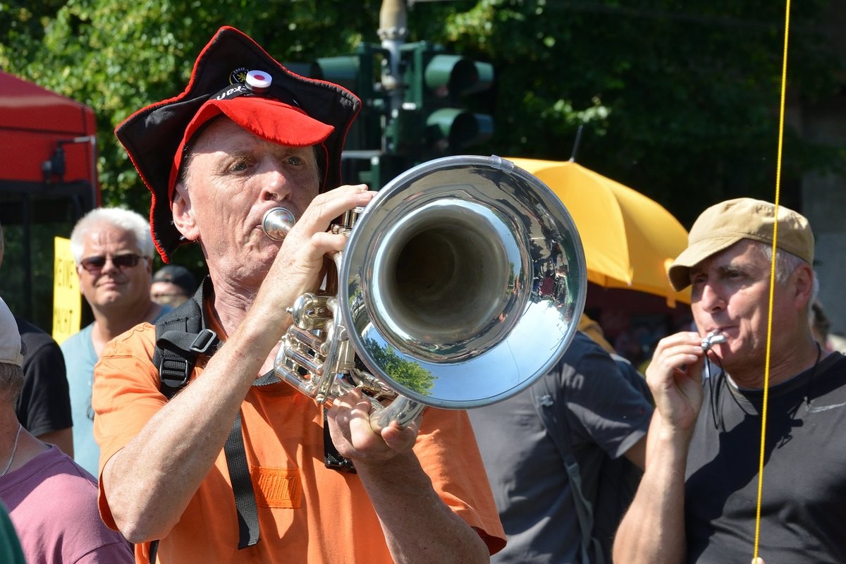 Ein Mann mit einer Art Narrenkappe spielt Trompete, daneben ein Mann mit Trillerpfeife im Demozug der Querdenker.