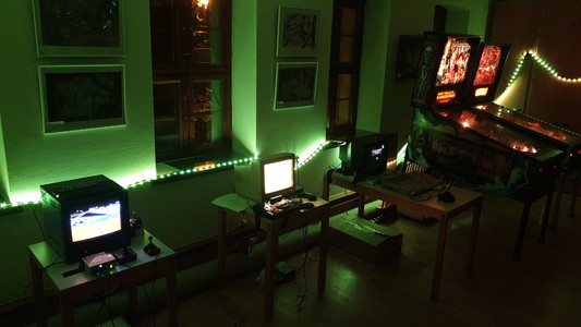 A row of three retro computers and two pinball machines in an illuminated room. From left side, green light.