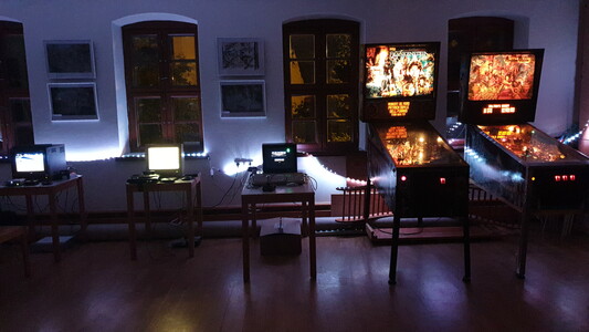 A row of three retro computers and two pinball machines in an illuminated room.
Front view, light blue illumination, windows behind.