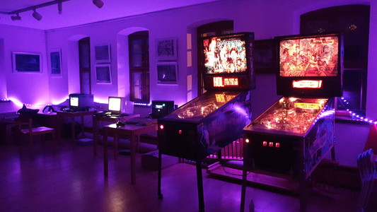 A row of three retro computers and two pinball machines in an illuminated room.
From right side, violet light.