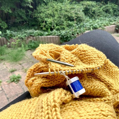 A view of a yellow knitted tee on the needles on a lap, with a slightly overgrown garden in the background 