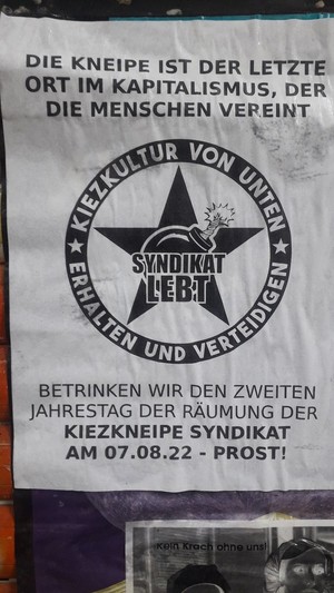Plakat mit der Aufschrift: Die Kneipe ist der letzte Ort im Kapitalismus, der die Menschen vereint! Betrinken wir den zweiten Jahrestag der Räumung der Kiezkneipe Syndikat  am 07.08.22 - Prost!