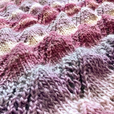 Close up of a knitted shawl in pink and lilac gradient yarn. The picture is taken under a slight angle, showing the hills and valleys that the fabric has due to its lace patterning.