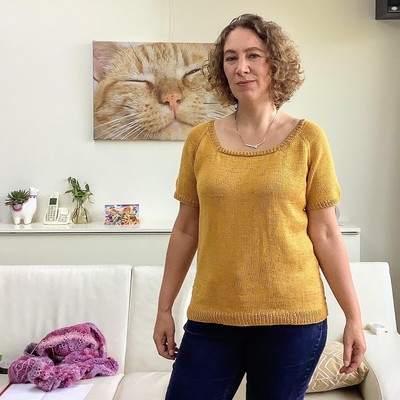 A picture a a woman with chin length curly hair, wearing a yellow knitted tee and blue jeans. She is looking into the camera, in front of a white couch and a wall with a close up picture of a red cat.