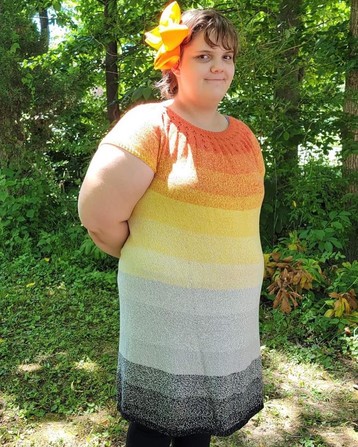 A girl with a large orange flower in  her hair wearing a knit dress in orange to black yarn. She is pictured from the front and looking into the camera 