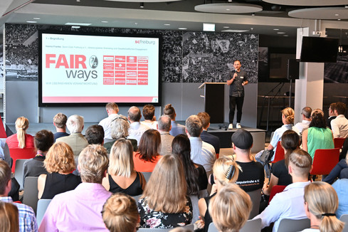 Tobias Rauber steht mit einem Mikrofon auf der Bühne. Neben ihm werden auf einer Leinwand die Logos der „FAIR ways“-Partner angezeigt. Im Vordergrund sieht man einen Teil des Publikums sitzen (Foto mit freundlicher Genehmigung des „SC Freiburg“).