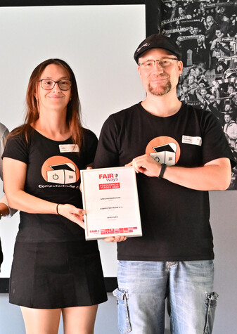 Julia Fiedler und Marco Rosenthal stehen lächelnd auf einer Bühne und halten gemeinsam eine gerahmte Urkunde in den Händen. Beide tragen ein schwarzes T-Shirt mit dem Logo der „Computertruhe“ (Foto mit freundlicher Genehmigung des „SC Freiburg“).