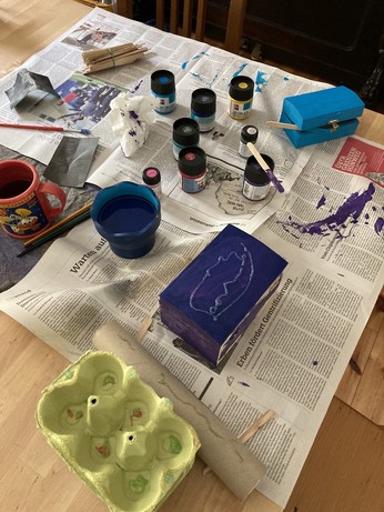 Übersichtsfoto: ein mit Zeitungspapier ausgelegter Tisch, einige Farbdöschen, Pinsel, zwei teilweise bemalte kleine Holzkästen in Form einer Schatzkiste, ein abstraktes Objekt aus Papprolle, Eierkartons, Glitzerklene