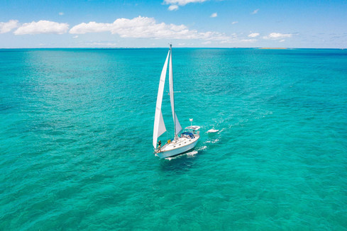 A sailboat in the middle of the ocean, moving forward with a jib and a mainsail on a bright sunny day. Green water, light winds, blue skies, nice white clouds on the horizon. (Source of the image: https://sailingsoulianis.com)