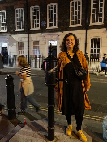 A woman smiling and standing next to a black water pump. The street seems busy with others walking in the background.