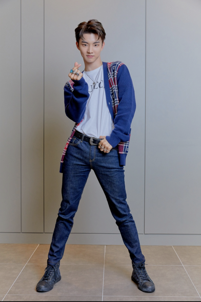 Ji Changmin wearing blue jeans, a blue cardigan with white and red plaid, a white GUCCI shirt, doing a heart sign.