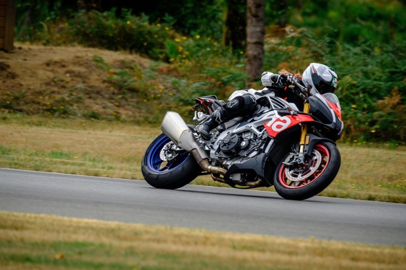 On track on my Aprilia Tuono Factory motorcycle. You can see green of grass and trees behind me, and my bike is mostly red, silver and black, with a blue rear wheel.
