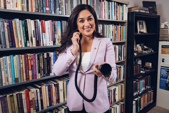 Photo of Jaipreet Virdi, a smiling South Asian woman with long hair. She is in an office, standing in front of a wall of bookshelves filled with books. She is wearing a lilac blazer and white blouse, and holding a conversation tube, with one end inserted in her ear, the other directed towards the viewer.
