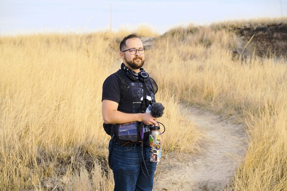 Tyler standing on a trail in Badlands National Park with field recording equipment including a microphone in his hands, a recorder, and headphones around his neck. The grass surrounding the trail is tall and light brown, like that commonly found a prairie environment. The trail leads up a small hill. The sky is on the horizon.
