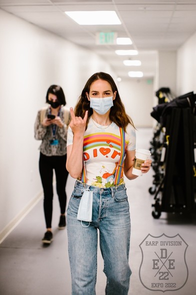 A photo of Wynonna Earp actress Melanie Scrofano doing a rocker hand gesture. She is wearing a rainbow Dior t-shirt and rainbow suspenders. 