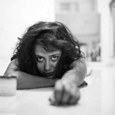Black and white image of Anu (moi) lying on the cold off-white marble floor with messy, wavy, untied, shoulder-length hair one hand stretched to the front and the other folded under my chin. There is a cup ashtray next to the folded hand. I am looking straight at the camera, eyes wide open, amused and almost smirking.
