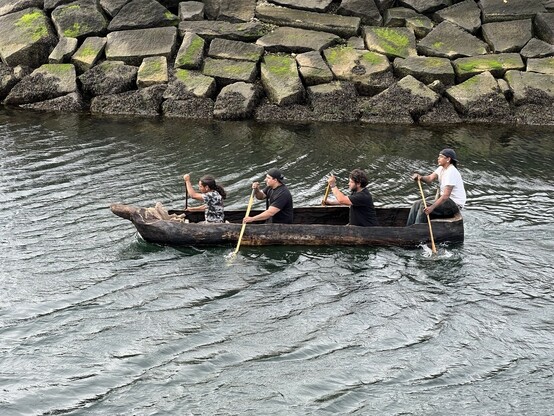 Group of four people paddle mishoon along slipway