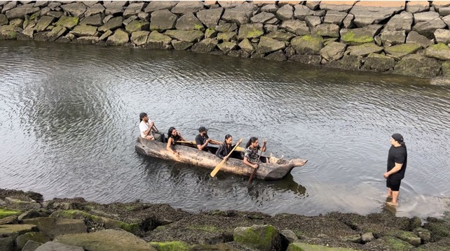 First group of 5 backing the mishoon (canoe) into the water with paddles for the first time while another person stands on shore having gotten them pushed off.