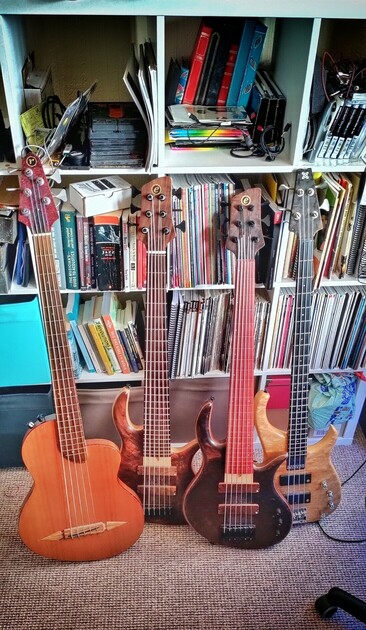 Photograph of 4 bass guitars leaning on a book case. Two have 6 strings, one has 5 strings and one has 4 strings. 