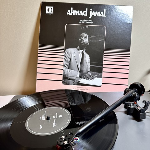 A record player with a black vinyl record. Tone arm across the record indicating it is halfway through side A. 

At the back, a pink record soeeve says: Ahmad Jamal, Live in Paris (1971), Lost ORTF Recordings