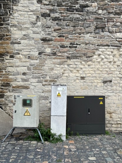 Three ugly electricity cabins against a very old stone wall.