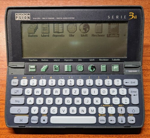 The "Serie 3a" (this is the German version) opened: Above the display: "16 bit CPU, Multi-Tasking, Digital Audio System". A dark green on lighter green non-backlit LC-Display sits above a shortcut bar "System" - "Daten" - "Word" - "Agenda" - "Uhr" - "Welt" - "Rechner" - "Tabelle". The keyboard below has nice rounded keys, large keys for Shift, Space, Enter and Backspace. 