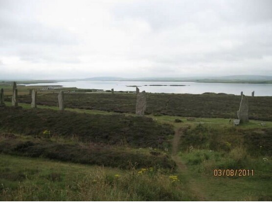 Ring of standing stones of which 12 can be seen. Outside the ring is a wide ditch & a higher mound. On the far side of the ring is Harray Lough which has some small islands in it. Land & mountains are in the distance. The sky is cloudy.