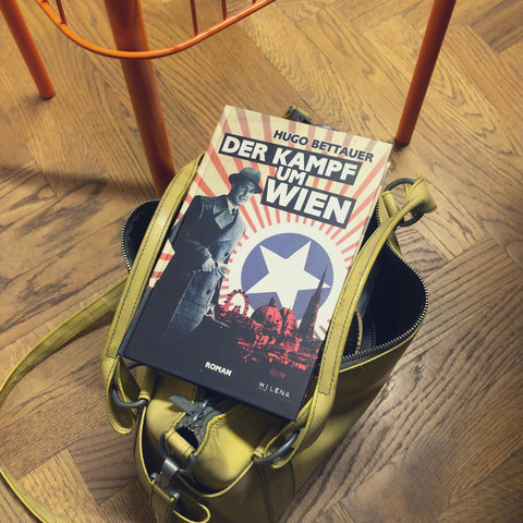 cover von hugo bettauers "der kampf um die stadt" aus gelber freytag-tasche schauend. die tasche steht auf einem holzfußboden neben einem orangen hocker.