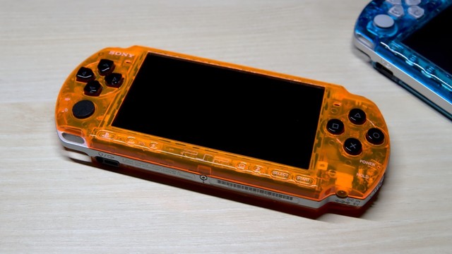 A PSP 3000 in a clear orange case, with black buttons. In the top right corner, parts of another PSP in a clear blue clase are visible.