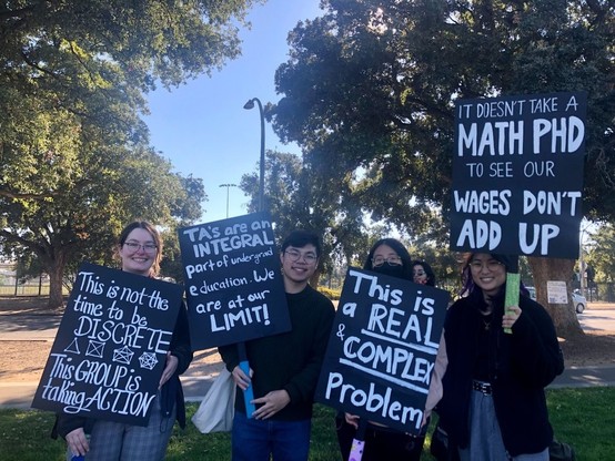 Signs say:
- "This is not the time to be DISCRETE. This GROUP is taking ACTION"
- "TAs are an INTEGRAL part of undergrad education. We are at our LIMIT!"
- "This is a REAL & COMPLEX Problem"
- "It doesn't take a math PhD to see our wages don't add up"