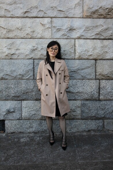 Korean woman in black glasses and a trenchcoat, standing in front of a gray stone wall