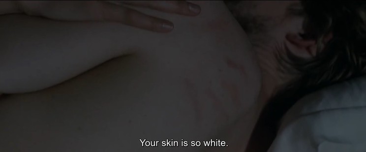Shot from the film Stars at Noon, showing the bare back of a white man in bed, shoulder blade blushed with a hand print from the white woman holding him, her hand on his arm. 'Your skin is so white.' reads the dialogue subtitle.