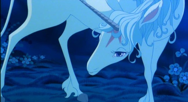 The unicorn bends low to the ground and looks into the camera, addressing a smaller creature. She is silver and white, but appears mostly blue in the shadows. A flower meadow can be seen faintly in the background.
