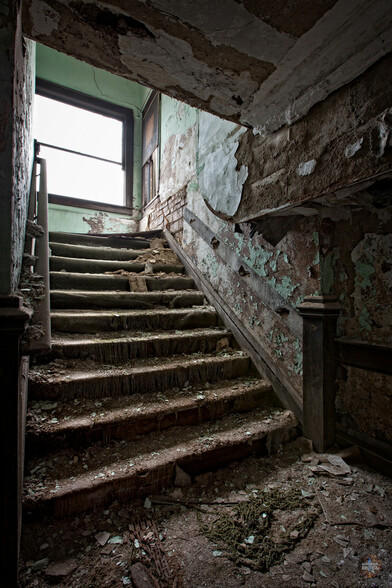 A rotting staircase leading up to a landing and window at the left. The walls are a light green that is flaking away to reveal plaster and brick underneath. The stairs are covered with carpet that has worn into individual strands hanging from each step. Paint flakes and debris from the walls and ceiling cover the stairs. 