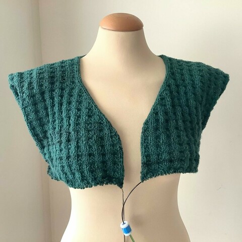 A cardigan-in-progress in petrol colored yarn and double broken rib patterning, is shown from the front on a mannequin against a white background. It has no sleeves yet, and just covers the bust.