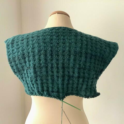 A cardigan-in-progress in petrol colored yarn and double broken rib patterning, is shown from the back on a mannequin against a white background. It has no sleeves yet.