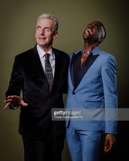 Peter Capaldi and Ncuti Gatwa in formal wear: Peter in a black suit and tie, Ncuti in an open-necked blue suit.  Peter is doing a Doctorish splayed hand and Ncuti has his head thrown back roaring with laughter.