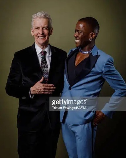 Peter Capaldi and Ncuti Gatwa in formal wear: Peter in a black suit and tie, Ncuti in an open-necked blue suit.  Ncuti is beaming at Peter, who is smiling at the camera.