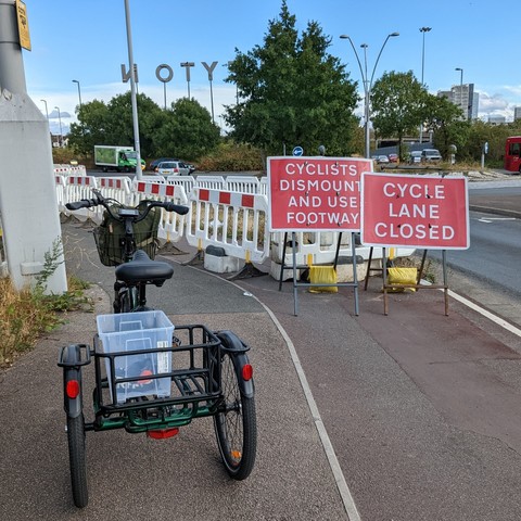 Photo of a trike stopped by a cycle lane with two signs. One says 'cycle lane closed' and the other says 'cyclists dismount and use footway'