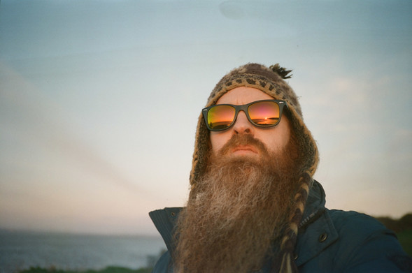 a man with a big ginger beard, wearing a brown hat and retro sunglasses with orange lenses. reflected in the lenses is an orange sunset and sea-scape.