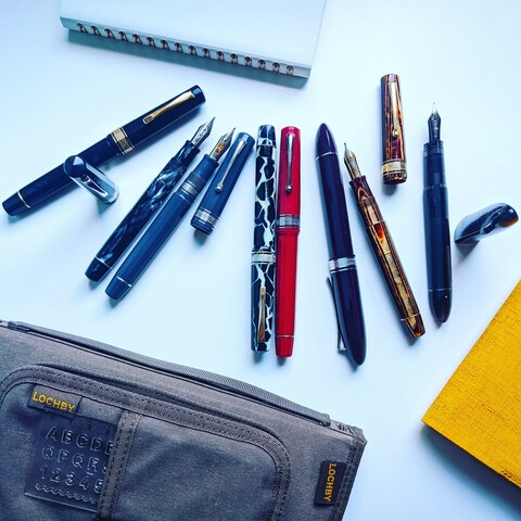 8 fountain pens on a white background with a pen pouch on top of a notebook at the bottom, a yellow notebook to the right and a white notebook in the top left corner.