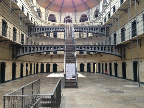 This is a photo of Kilmainham Gaol, a former prison in Kilmainham, Dublin, Ireland. It was built in 1796. The photo shows a large open hall with three floors of prison cells accessed by a central stairway, which is in the middle of the photo.