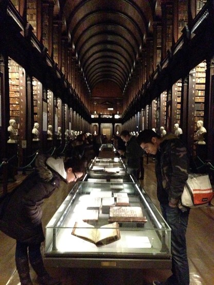 This is a photo of Trinity College Library in Dublin, Ireland. It shows a long room with a long glass case flanked by alcoves with shelves of old books and white busts of significant historical figures.